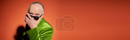 älterer grauhaariger Mann in grünem Velours-Blazer, der in die Kamera blickt, über dunkler und trendiger Sonnenbrille auf rotem und orangefarbenem Hintergrund, älteres Modell, positives und stilvolles Alterskonzept, Banner mit Kopierraum