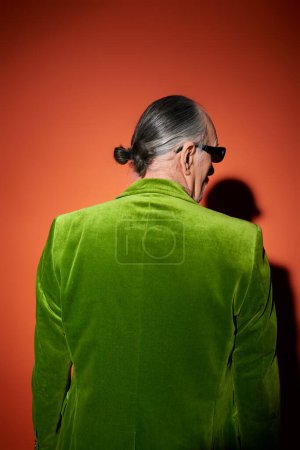 Rückansicht eines grauhaarigen älteren Mannes in trendiger Freizeitkleidung, grünem Velours-Blazer und dunkler Sonnenbrille, der auf rotem und orangefarbenem Hintergrund mit Schatten steht, modisches älteres männliches Modell