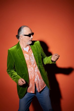 cool senior man in dark sunglasses, green velour blazer and trendy shirt having fun, dancing and looking away on red and orange background with shadow, vibrant personality, happy aging concept