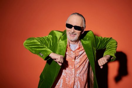 joy and happiness, excited and fashionable senior man in dark sunglasses, trendy shirt and green velour blazer having fun and dancing on red and orange background with shadow