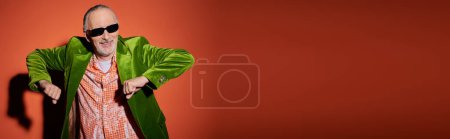 cheerful elderly man in dark sunglasses, green velour blazer and trendy shirt dancing and having fun on red and orange background with shadow, vibrant personality, banner with copy space