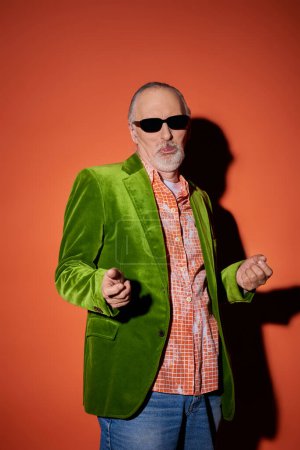 cool senior man in dark sunglasses, trendy shirt and green velour blazer gesturing and pouting lips while having fun and posing on red and orange background with shadow, fashionable and happy aging