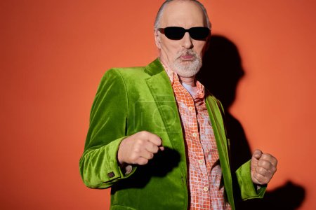 Cooler Senior mit dunkler Sonnenbrille, trendigem Hemd und grünem Velours-Blazer, schmollenden Lippen und tanzendem Spaß auf rotem und orangefarbenem Hintergrund mit Schatten, Happy-Aging-Konzept