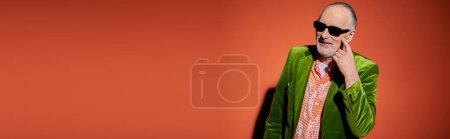 fröhlicher älterer Herr mit dunkler Sonnenbrille, schickem Hemd und grünem Velours-Blazer, der auf die Wange zeigt und um einen Kuss auf rotem und orangefarbenem Hintergrund bittet, glückliches Altern, Banner mit Kopierplatz