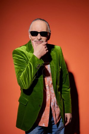 uśmiechnięty starszy mężczyzna w ciemnych okularach i zielonym welurowym blezerze dotykający brody i patrzący w kamerę na czerwono-pomarańczowym tle, osobisty styl, koncepcja szczęśliwego starzenia się