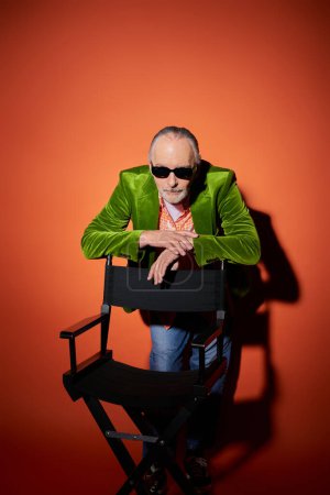 personnalité senior vibrante et élégante, homme âgé aux lunettes de soleil sombres et au blazer en velours vert appuyé sur une chaise et regardant la caméra sur fond rouge et orange avec ombre, concept de vieillissement positif