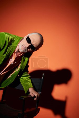 Foto de Hombre anciano reflexivo y curioso en gafas de sol oscuras y chaqueta de terciopelo verde de pie cerca de la silla y mirando hacia otro lado en el fondo rojo y naranja con sombra, envejecimiento de moda, estilo de vida elegante - Imagen libre de derechos