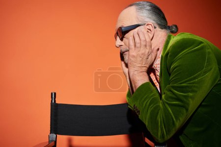 Foto de Vista lateral del modelo de pelo gris senior tocando la cara y mirando hacia otro lado cerca de la silla sobre fondo rojo y naranja, look de moda, gafas de sol oscuras, chaqueta de terciopelo verde, concepto de envejecimiento de moda - Imagen libre de derechos