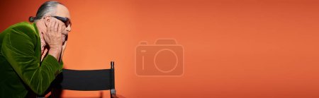 Foto de Vista lateral del modelo masculino senior en chaqueta de terciopelo verde y gafas de sol oscuras de pie cerca de la silla, tocando la cara y pensando en el fondo rojo y naranja, estilo personal, moda casual, pancarta - Imagen libre de derechos