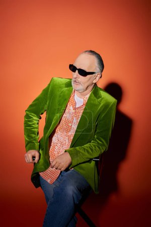 älterer und selbstbewusster bärtiger Mann auf Stuhl sitzend und auf rotem und orangefarbenem Hintergrund mit Schatten wegschauend, Modelook, dunkle Sonnenbrille, trendiges Hemd, grüner Velours-Blazer, Mode- und Alterskonzept