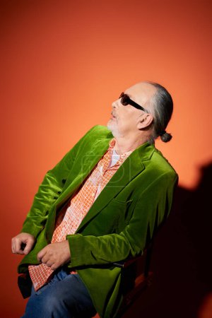 hombre mayor sorprendido en gafas de sol oscuras, camisa de moda y chaqueta de terciopelo verde sentado en la silla y mirando hacia otro lado en el fondo rojo y naranja con sombra, look de moda, concepto de envejecimiento positivo