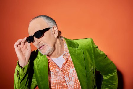 individualidad vibrante, concepto de moda y edad, hombre barbudo senior con camisa de moda y chaqueta de terciopelo verde ajustando las gafas de sol oscuras mientras está sentado y mirando hacia otro lado en el fondo rojo y naranja