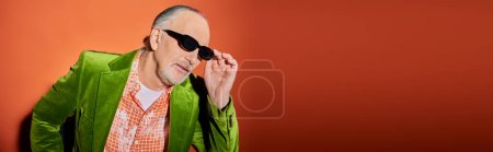 senior man model in trendy shirt and green velour blazer touching dark sunglasses and looking away on red and orange background, stylish casual clothes, fashion and age concept, banner