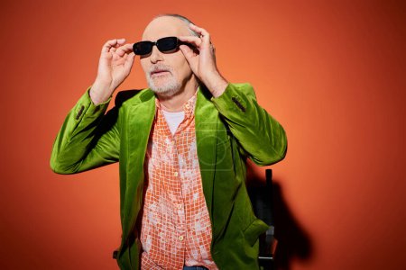 Positives und modisches Alterskonzept, älteres männliches Modell in grünem Velours-Blazer und trendigem Hemd, das vom Stuhl auf rotem und orangefarbenem Hintergrund wegschaut und eine dunkle Sonnenbrille aufsetzt