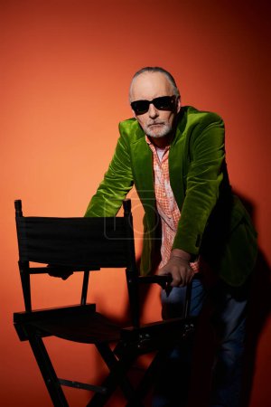 hombre mayor seguro y serio en gafas de sol oscuras, camisa de moda y chaqueta de terciopelo verde de pie cerca de la silla y mirando a la cámara en el fondo rojo y naranja con sombra, estilo personal