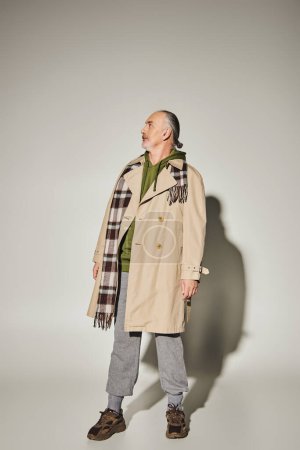 full length of senior male model in beige trench coat, plaid scarf and green hoodie standing on grey background with shadow and looking away, stylish casual attire, fashionable aging concept