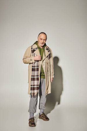 fashion look, positive aging, full length of smiling senior male model in green hoodie, beige trench coat and plaid scarf standing and looking at camera on grey background with shadow