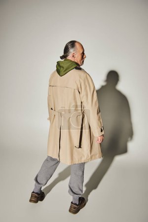 vue arrière du mannequin senior en vêtements décontractés élégants debout sur fond gris avec ombre, homme âgé et aux cheveux gris en trench-coat beige et sweat à capuche vert, concept de mode de vie tendance