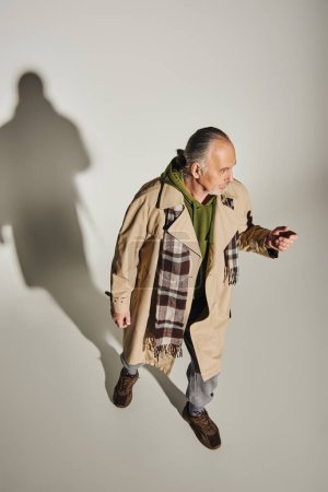 longueur totale de l'homme âgé en tenue décontractée élégant debout sur fond gris avec ombre et détournement des yeux, sweat à capuche vert, trench coat beige, écharpe à carreaux, concept de vieillissement à la mode, vue grand angle