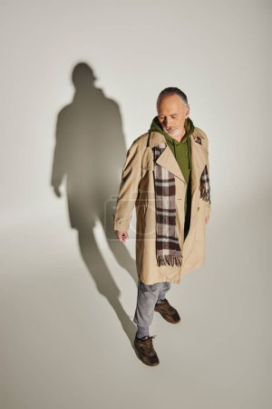 homme âgé et barbu en trench-coat beige, sweat à capuche vert et foulard à carreaux debout sur fond gris avec ombre, tenue décontractée tendance, style personnel, vue grand angle
