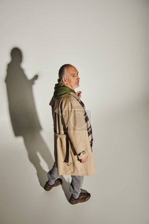 full length of thoughtful senior man in green hoodie, beige trench coat and plaid blazer looking away on grey background with shadow, fashionable lifestyle, high angle view