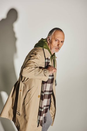 confident senior man with serious face expression standing and looking at camera on grey background with shadow, casual attire, plaid scarf, beige trench coat, green hoodie, stylish older model