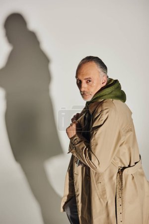 Mode und Alter, persönlicher Stil, selbstbewusster älterer Herr mit ernstem Gesichtsausdruck, der im beigen Trenchcoat steht und auf grauem Hintergrund mit Schatten in die Kamera schaut