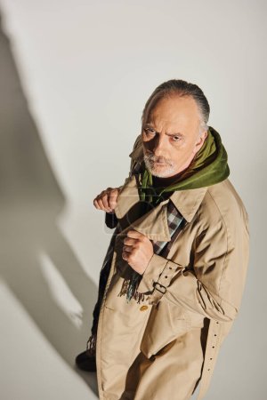 high angle view of serious and strict senior man looking at camera when standing on grey background with shadow, green hoodie, beige trench coat, plaid scarf, expressive gaze, trendy aging concept