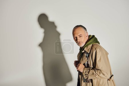 Photo for Senior and thoughtful male model in green hoodie, plaid scarf and beige trench coat standing on grey background with shadow and looking away, personal style, fashion and age concept - Royalty Free Image