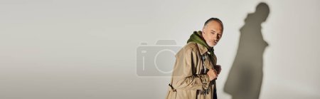 fashion and age concept, pensive senior man in green hoodie, beige trench coat and plaid scarf looking away while standing on grey background with shadow, banner with copy space