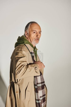 grey haired, bearded and pensive senior man posing in stylish casual clothes on grey background, beige trench coat, green hoodie, plaid scarf, fashion and age, aging population concept