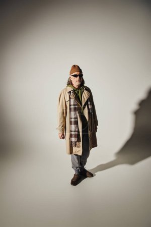 Mode- und Alterskonzept, hipsterhafter Senior mit Hut, dunkler Sonnenbrille, beigem Trenchcoat und kariertem Schal auf grauem Hintergrund mit Schatten und Kopierraum