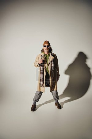 Photo for Senior hipster, full length of trendy aged man in dark sunglasses, beanie hat, beige trench coat and plaid scarf standing on grey background with shadow, fashionable aging concept - Royalty Free Image