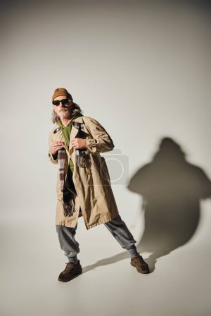 Téléchargez les photos : Personnalité expressive et à la mode, pleine longueur de l'homme âgé en vêtements de style hipster et lunettes de soleil sombres posant sur fond gris avec ombre, concept de style de vie positif - en image libre de droit