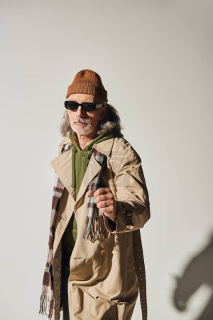 Téléchargez les photos : Homme âgé confiant dans des lunettes de soleil sombres et tenue de style hipster debout avec poing serré et regardant la caméra sur fond gris, bonnet chapeau, trench coat beige, concept de vieillissement à la mode - en image libre de droit