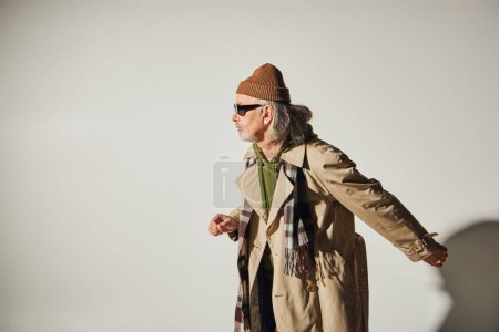 Téléchargez les photos : Vue latérale de l'homme âgé en vêtements décontractés à la mode marchant sur fond gris, bonnet chapeau, lunettes de soleil sombres, trench coat beige, écharpe à carreaux, style hipster, concept de vieillissement à la mode - en image libre de droit