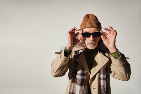 envejecimiento de moda, estilo hipster, hombre mayor fresco en sombrero de gorro, bufanda a cuadros y gabardina ajustando gafas de sol oscuras y mirando a la cámara en el fondo gris, sesión de fotos de moda