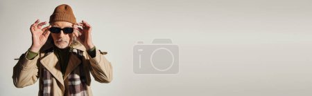 Téléchargez les photos : Cool senior homme en bonnet chapeau, trench coat beige et foulard à carreaux ajuster les lunettes de soleil à la mode et en regardant la caméra sur fond gris, mode hipster, vieillissement élégant, bannière avec espace de copie - en image libre de droit