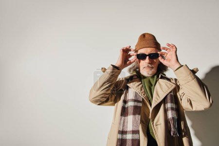 homme âgé barbu en bonnet chapeau, écharpe à carreaux et élégant trench coat regardant la caméra tout en se tenant sur fond gris, style hipster, individualité expressive, concept de vieillissement à la mode