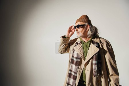 anciano en gorro sombrero, gabardina beige y bufanda a cuadros ajustando gafas de sol oscuras y mirando hacia otro lado sobre fondo gris, estilo hipster, individualidad, moda y concepto de edad