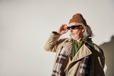 expressive personality, hipster style senior man in beanie hat, beige trench coat and plaid scarf adjusting dark sunglasses and looking away while standing on grey background