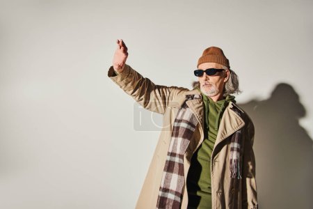hombre mayor de moda en gafas de sol oscuras, gorro sombrero, gabardina beige y bufanda a cuadros de pie con extendido y mirando hacia otro lado sobre fondo gris, estilo hipster, personalidad expresiva