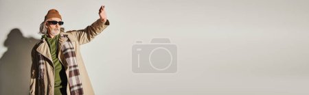 Photo for Senior hipster style man in trendy casual clothes and dark sunglasses standing with outstretched hand and looking away on grey background, fashionable aging concept, banner with copy space - Royalty Free Image