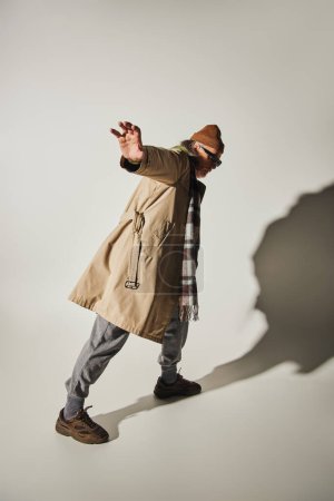longitud completa del hombre hipster senior mostrando gesto de stop mientras está de pie en postura expresiva sobre fondo gris con sombra, gafas de sol oscuras, gorro sombrero, gabardina beige, moda y concepto de edad