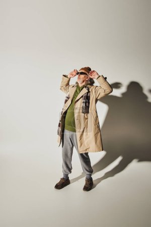 Téléchargez les photos : Longueur totale de cool senior homme en bonnet chapeau et trench coat beige ajuster les lunettes de soleil sombres et regarder loin sur fond gris avec ombre, tenue décontractée de style hipster, concept de mode et d'âge - en image libre de droit