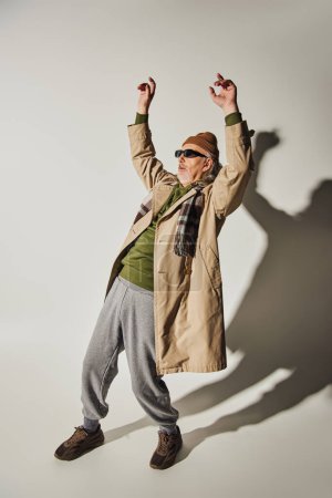 volle Länge des ausdrucksstarken Hipster-Stils Senior mit dunkler Sonnenbrille, Beanie-Hut, beigem Trenchcoat und kariertem Schal posiert mit erhobenen Händen auf grauem Hintergrund mit Schatten, Tanz