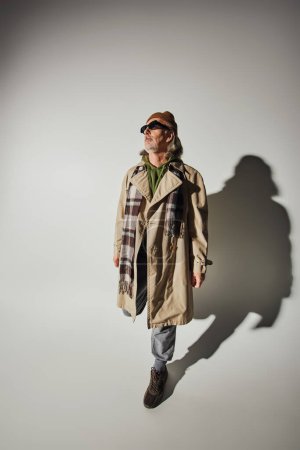 Hipster-Stil, modisches alterndes Konzept, hochaufragende Ansicht eines älteren Mannes mit Hut, dunkler Sonnenbrille, beigem Trenchcoat und kariertem Schal, der auf grauem Hintergrund mit Schatten posiert