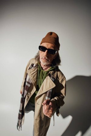 Photo for Hipster style, trendy casual clothes, senior man in dark sunglasses, beanie hat and plaid scarf looking at camera on grey background with shadow, fashionable lifestyle and positive aging concept - Royalty Free Image