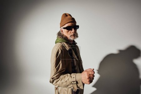 älteres männliches Model mit dunkler Sonnenbrille und stylischer Freizeitkleidung, die auf grauem Hintergrund mit Schatten in die Kamera blickt, Mütze, beiger Trenchcoat, karierter Schal, ausdrucksstarke Persönlichkeit