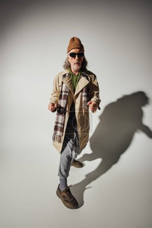 longueur complète de l'homme âgé à la mode et cool dans des lunettes de soleil sombres, bonnet chapeau et beige trench coat pointant avec les doigts à la caméra sur fond gris avec ombre, modèle senior de style hipster